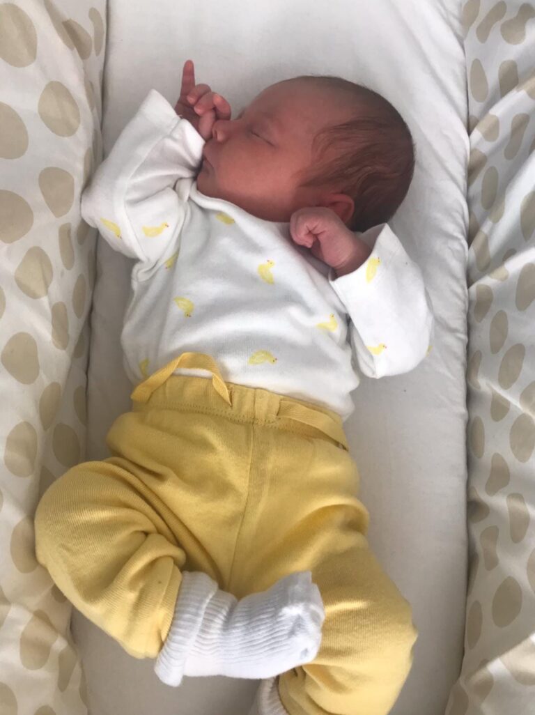 A newborn baby wearing yellow trousers, stretching and reaching out with their arms in a peaceful, natural pose.